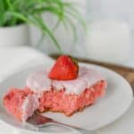 A slice with pink frosting and a whole piece of fruit on top, served on a white plate with a fork.