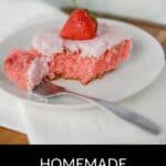 A slice of homemade strawberry cake with pink frosting and a strawberry on top sits on a white plate with a fork, resting on a white napkin.
