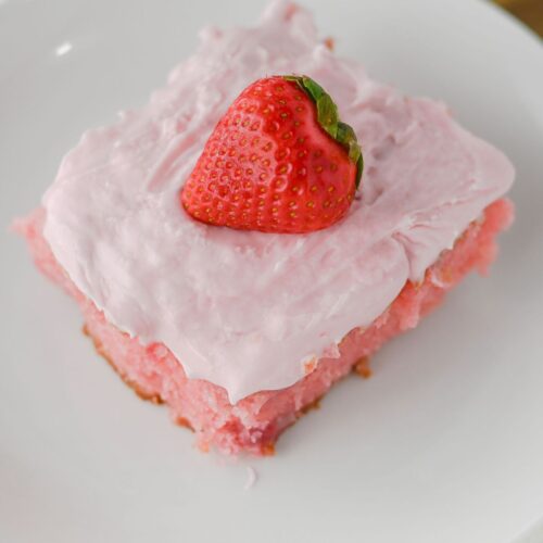 A square slice of homemade strawberry cake with pink frosting, topped with a whole strawberry, served on a white plate.