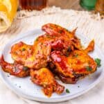 Plate of barbecued chicken pieces, including tender slow cooker chicken wings, garnished with chopped herbs and served on a white plate with bread and drinks in the background.