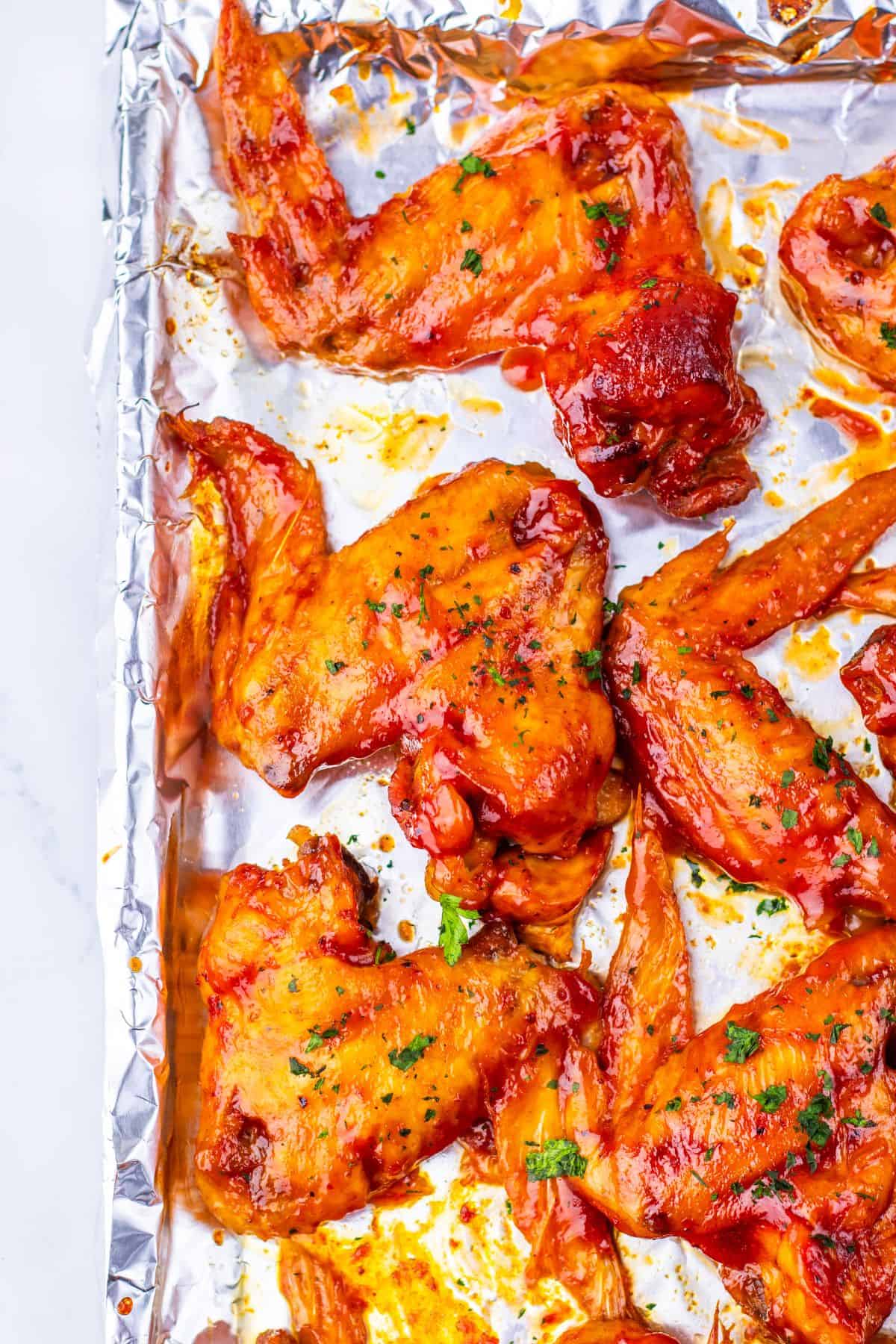 Slow Cooker Chicken Wings baked and coated in a zesty red sauce are arranged on a foil-lined baking sheet, then garnished with fresh chopped herbs.