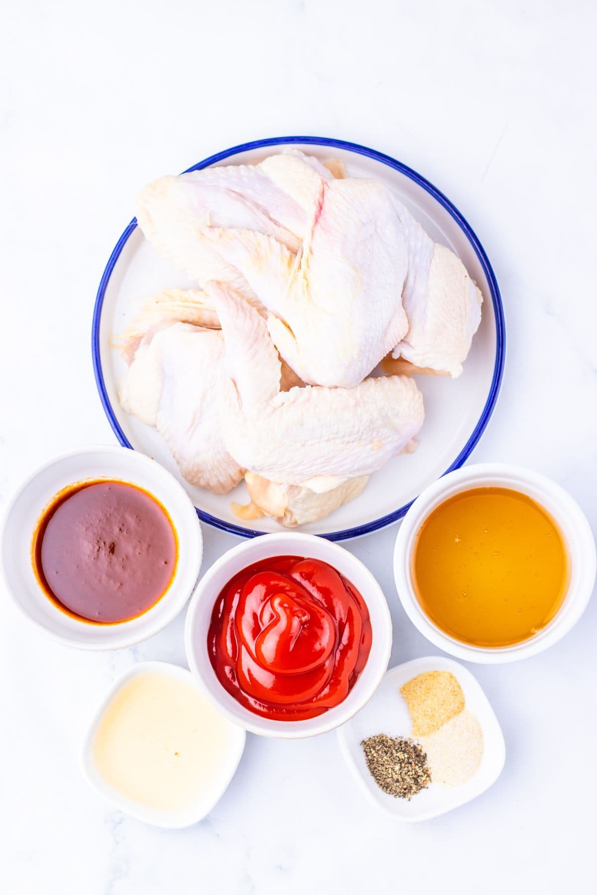 A plate of raw Slow Cooker Chicken Wings is surrounded by bowls of barbecue sauce, ketchup, honey, mayonnaise, and small dishes of spices on a white surface.