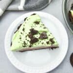 A slice of Oreo Pudding Pie with light green topping and chocolate cookie crumbs sits on a white plate, with cookies and a striped cloth nearby.