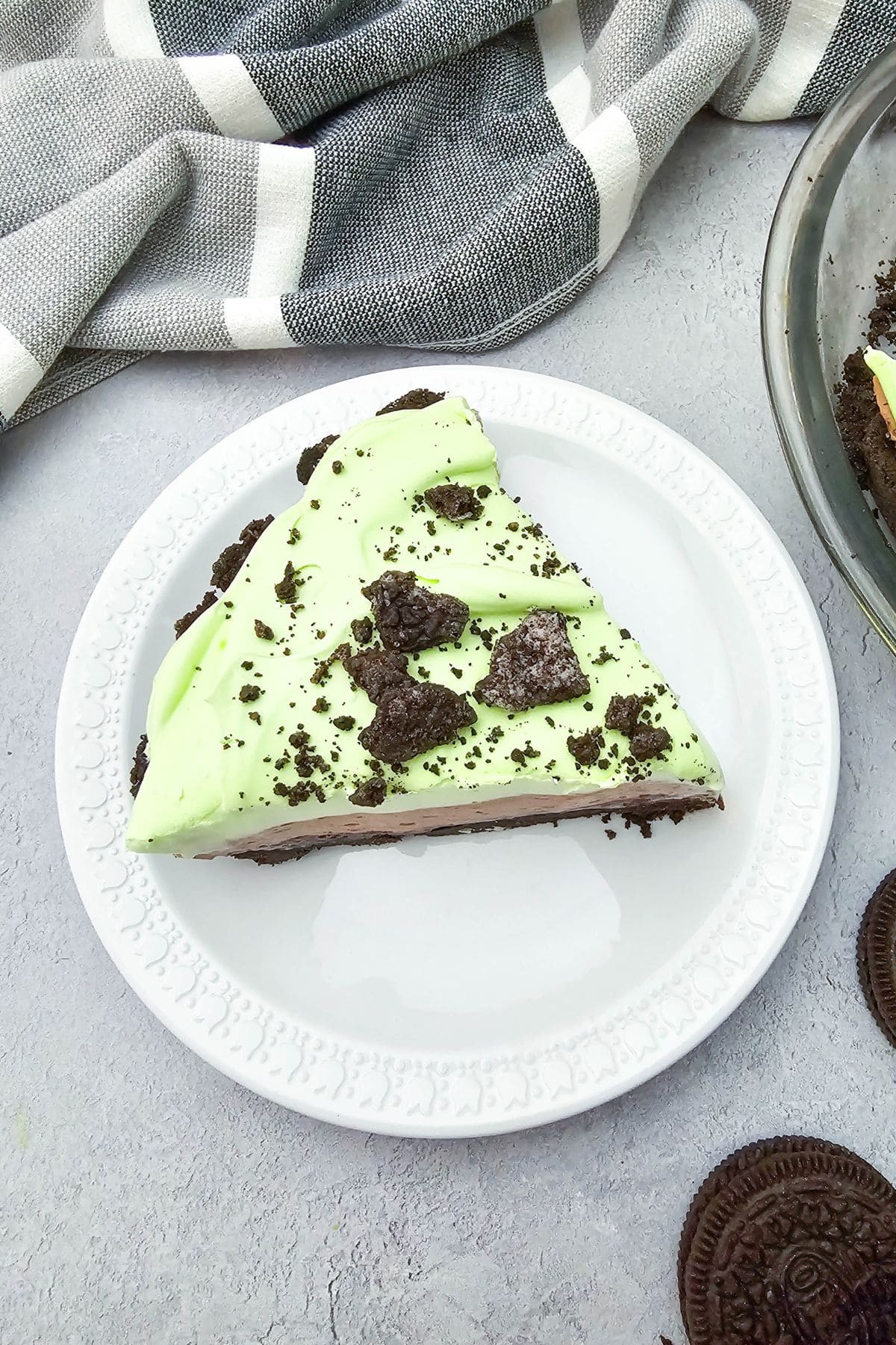 A slice of Oreo Pudding Pie with light green topping and chocolate cookie crumbs sits on a white plate, with cookies and a striped cloth nearby.