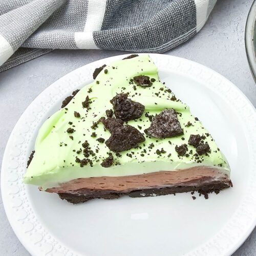 A slice topped with crushed cookies, sits on a white plate atop a gray surface with a checkered cloth nearby.