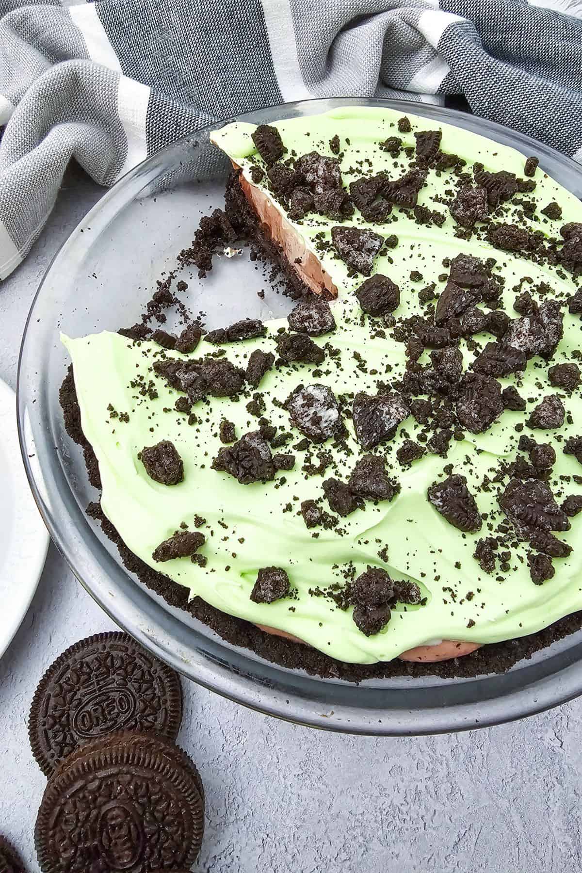 Green cream filling and chocolate cookie crust sits in a glass dish, topped with crumbled cookies. A slice is missing. Two  are on the table nearby.
