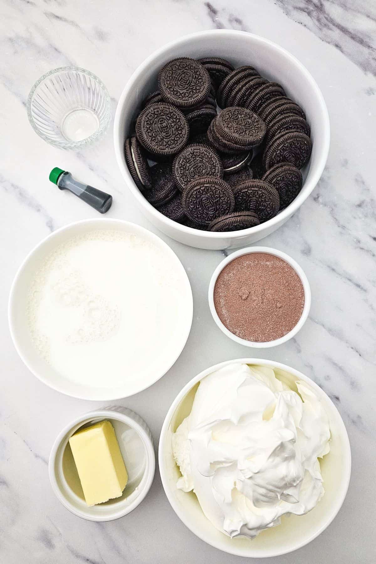 Ingredients on a marble surface: Cookies, cocoa powder, whipped cream, butter, milk, green food coloring, and a small glass bowl.
