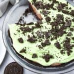 A glass pie dish with a partially sliced Oreo Pudding Pie, topped with green cream and crumbled Oreo cookies. Two Oreo cookies are on the table nearby.