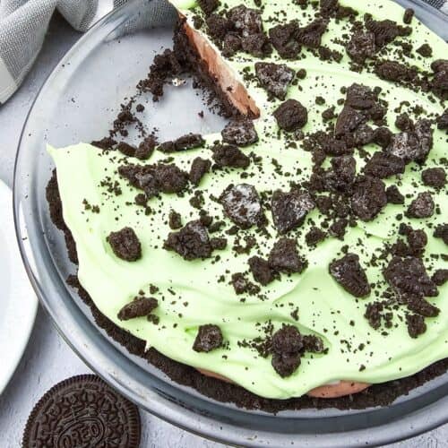 A glass pie dish with a partially sliced Oreo Pudding Pie, topped with green cream and crumbled Oreo cookies. Two Oreo cookies are on the table nearby.