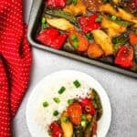 A plate of Sheet Pan Chicken with Vegetables—white rice, chicken, snap peas, red bell peppers, and scallions—sits beside a baking tray of the same dish. A red polka-dot cloth rests next to the tray.