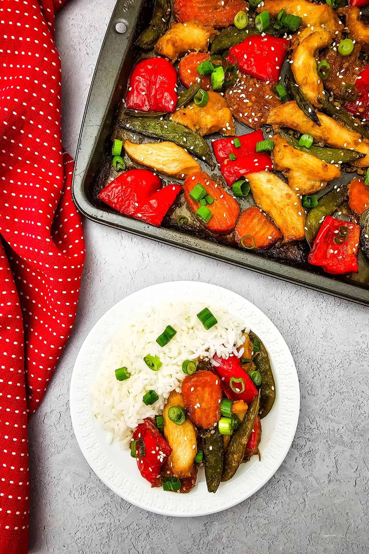 A plate of Sheet Pan Chicken with Vegetables—white rice, chicken, snap peas, red bell peppers, and scallions—sits beside a baking tray of the same dish. A red polka-dot cloth rests next to the tray.