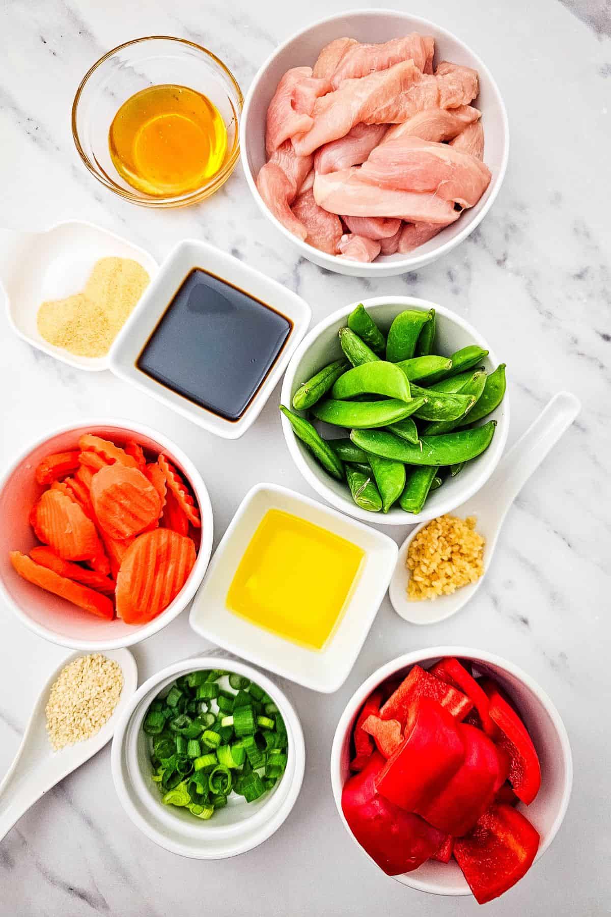 A variety of fresh ingredients in bowls, including sliced raw chicken for Sheet Pan Chicken with Vegetables, snap peas, sliced carrots, red bell peppers, green onions, minced garlic, soy sauce, oil, and seasonings.