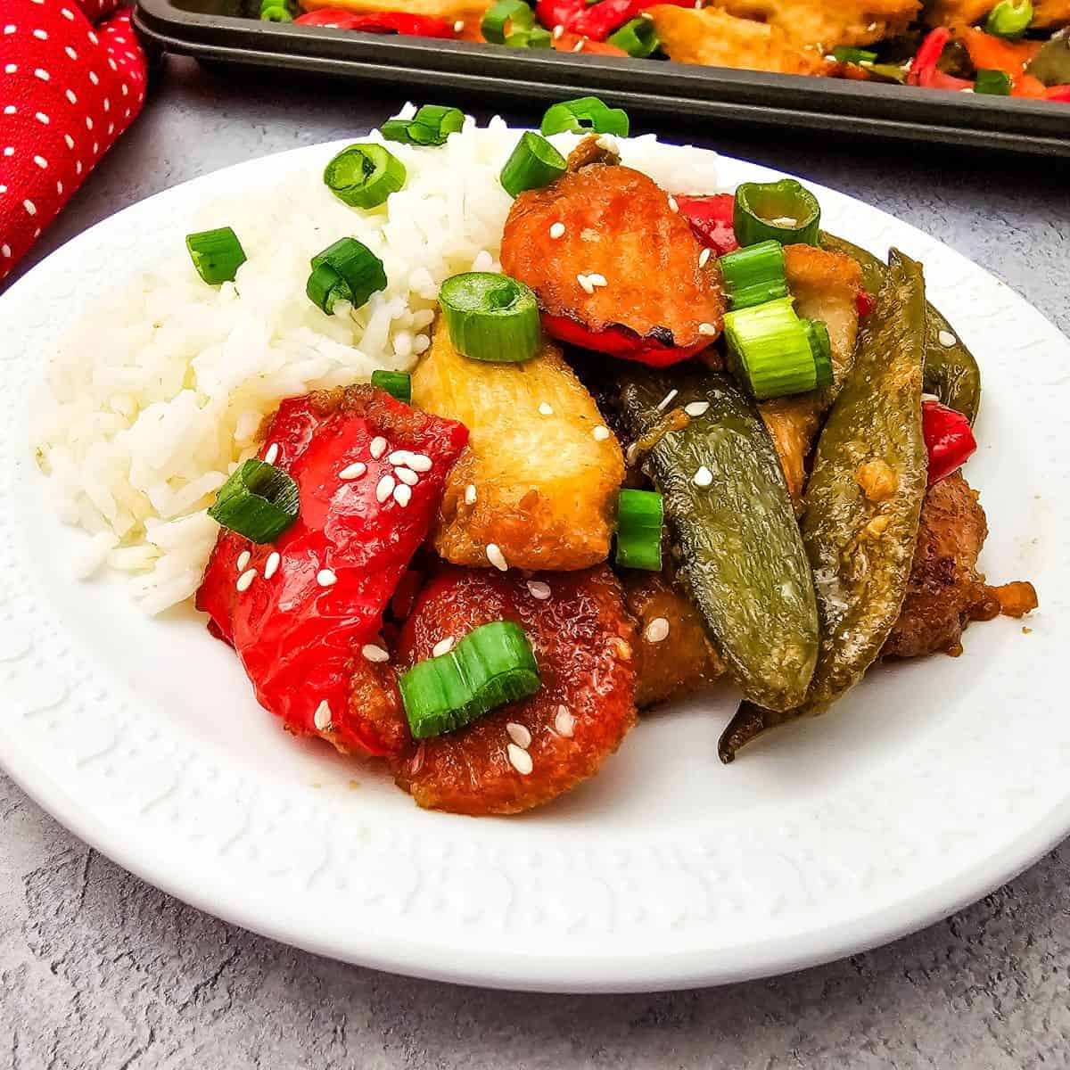 A plate of white rice served with Sheet Pan Chicken with Vegetables—tender chicken, red bell peppers, carrots, snap peas—topped with chopped green onions and sesame seeds.