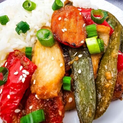 A plate of white rice served with Sheet Pan Chicken with Vegetables, featuring stir-fried chicken, red bell peppers, snap peas, chopped green onions, and sesame seeds.