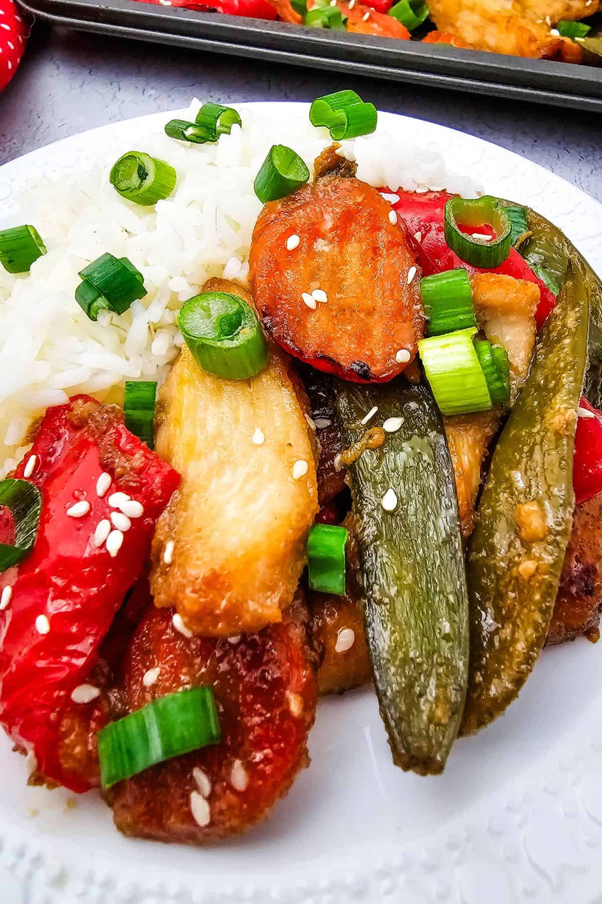 A plate of white rice served with Sheet Pan Chicken with Vegetables, featuring stir-fried chicken, red bell peppers, snap peas, chopped green onions, and sesame seeds.