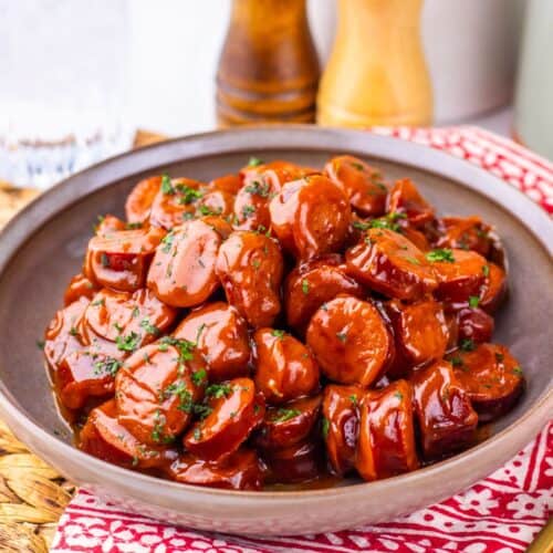 A bowl of sliced Crock Pot Kielbasa coated in barbecue sauce, garnished with herbs, sits on a red-patterned napkin with pepper and salt mills in the background.