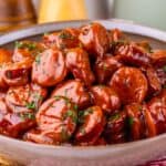 A bowl filled with sliced Crock Pot Kielbasa coated in glossy barbecue sauce, garnished with chopped herbs, sits on a red-patterned cloth. Salt and pepper shakers are blurred in the background.