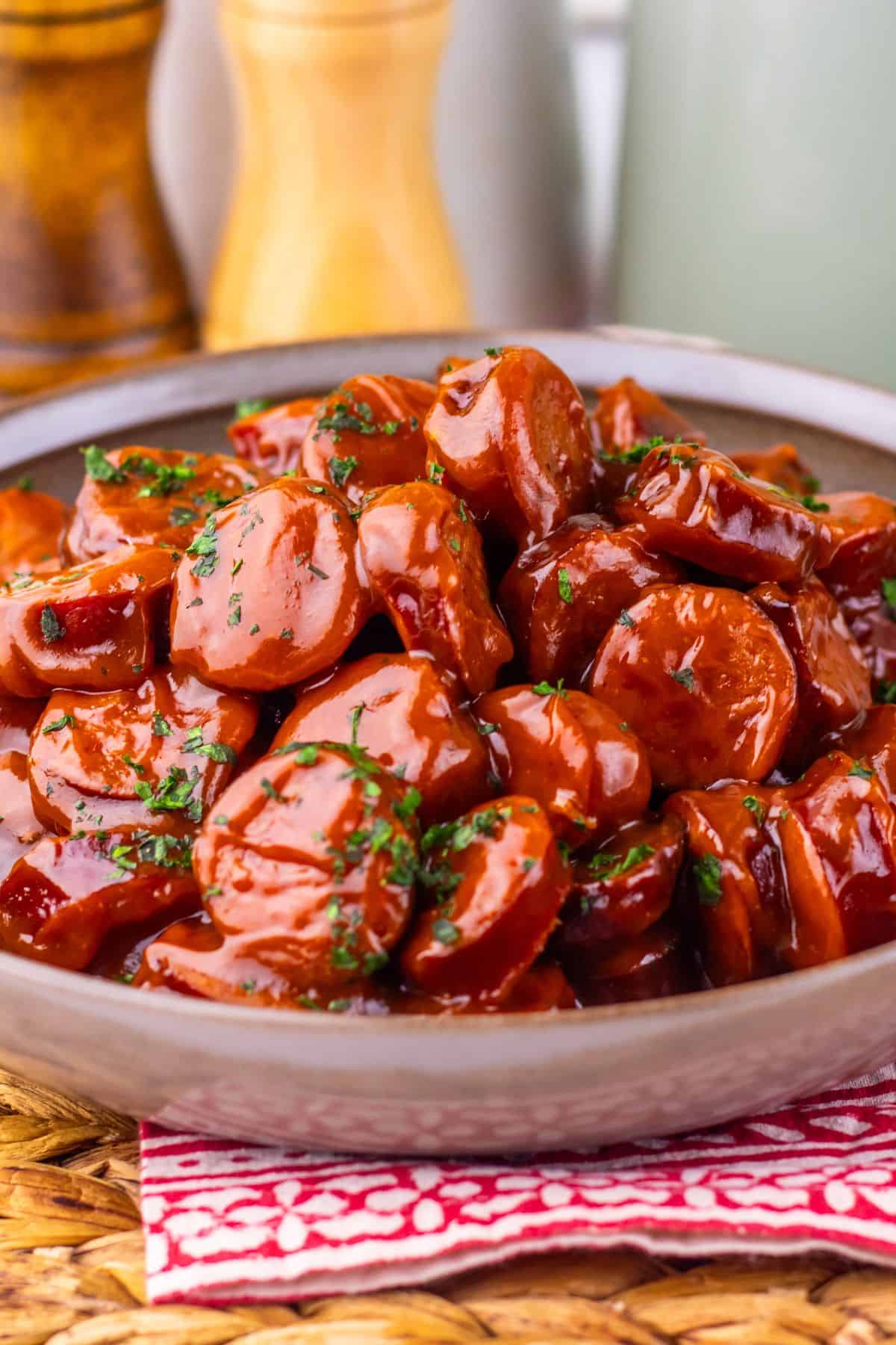 A bowl filled with sliced Crock Pot Kielbasa coated in glossy barbecue sauce, garnished with chopped herbs, sits on a red-patterned cloth. Salt and pepper shakers are blurred in the background.