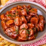 A plate of meat in a glossy brown sauce, garnished with chopped herbs, sits on a brown dish atop a woven placemat with a red and white napkin underneath.