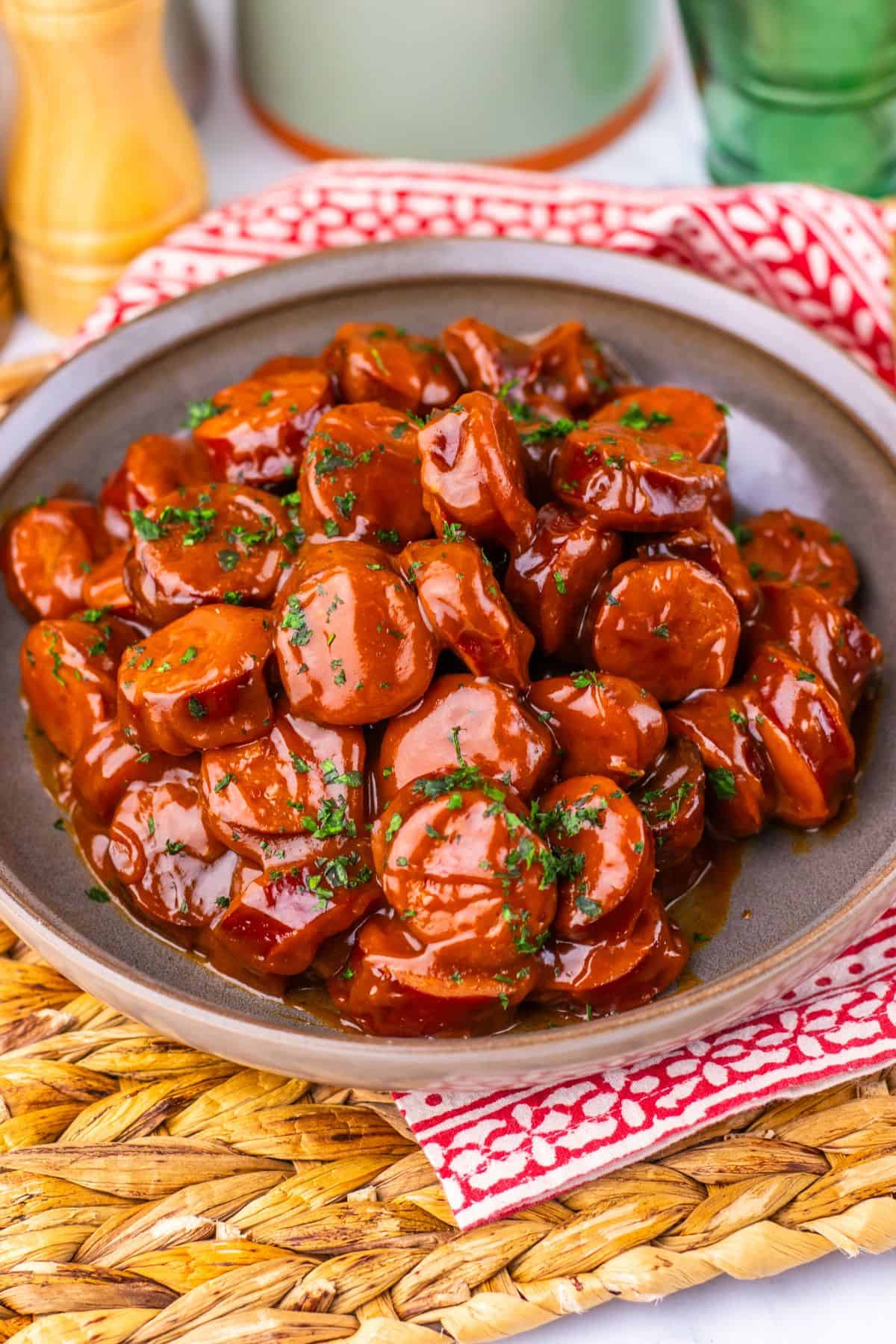 A plate of meat in a glossy brown sauce, garnished with chopped herbs, sits on a brown dish atop a woven placemat with a red and white napkin underneath.