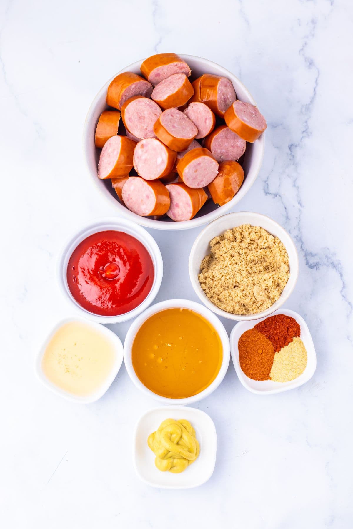 A bowl of sliced Crock Pot Kielbasa is surrounded by small bowls containing ketchup, brown sugar, honey, mustard, vinegar, and assorted spices on a white marble surface.