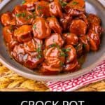 A plate of sliced Crock Pot Kielbasa sausage bites coated in sauce, garnished with herbs, sits on a gray plate with a red and white cloth underneath. The text reads: "Crock Pot Kielbasa Bites.