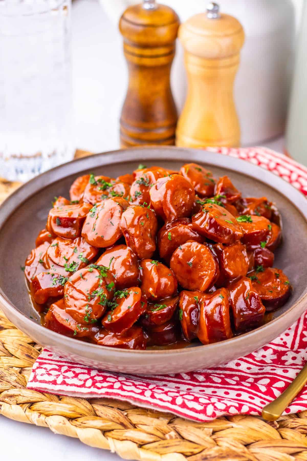 A bowl of Crock Pot Kielbasa in a glossy brown sauce, garnished with herbs, sits on a red patterned napkin with salt and pepper shakers in the background.