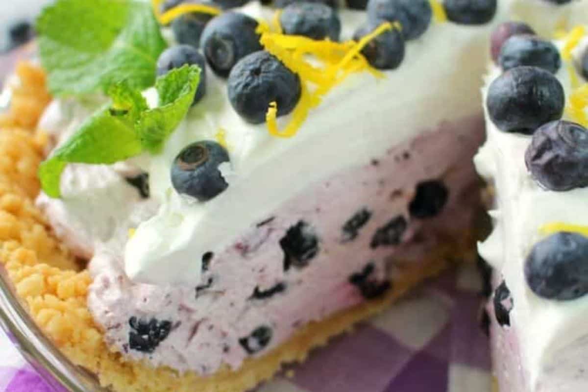 A close-up of a blueberry cream pie with a crumb crust, topped with whipped cream, fresh blueberries, lemon zest, and a mint sprig. One slice has been removed from this spring dessert—one of the prettiest desserts for seasonal celebrations.