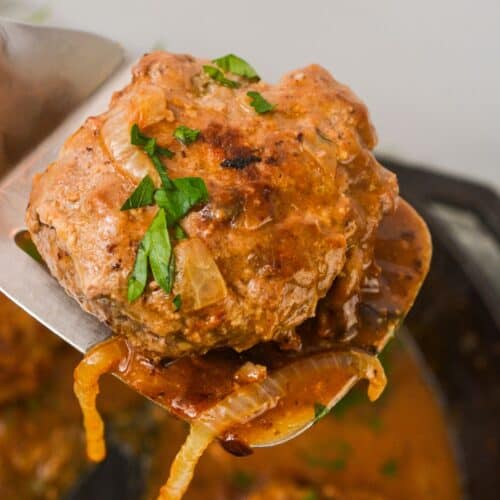 A cooked Hamburger Steak with Onion Gravy is lifted on a metal spatula, garnished with chopped parsley.