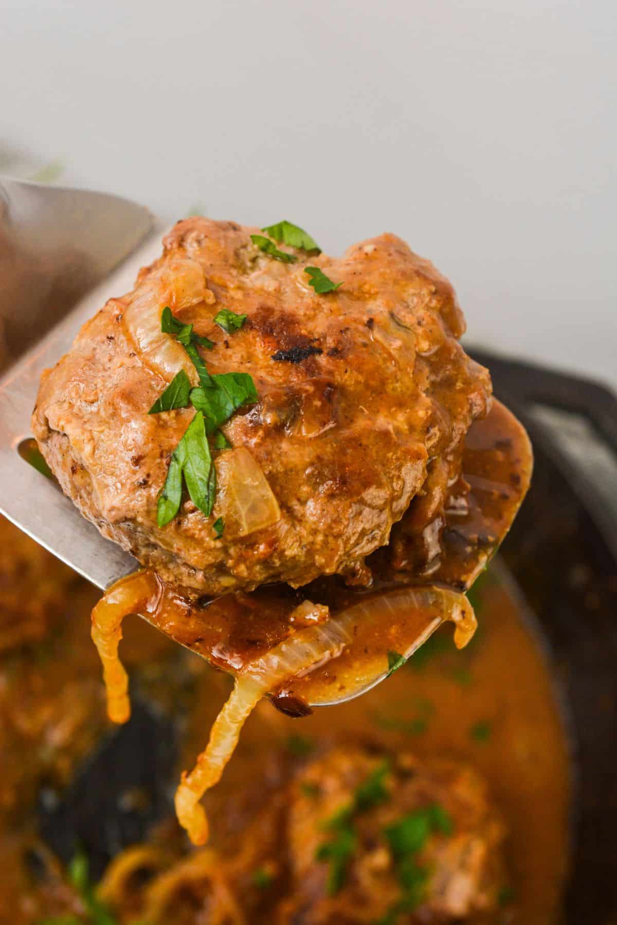 A cooked Hamburger Steak with Onion Gravy is lifted on a metal spatula, garnished with chopped parsley.