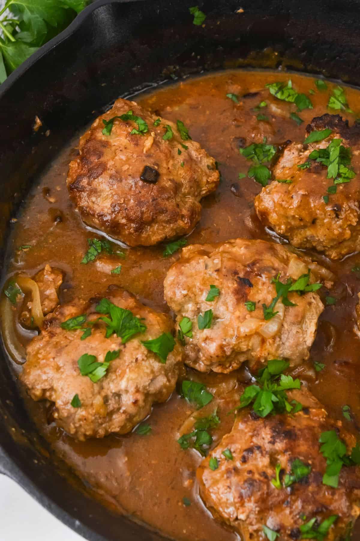 Five browned meat patties simmer in a skillet garnished with chopped parsley.