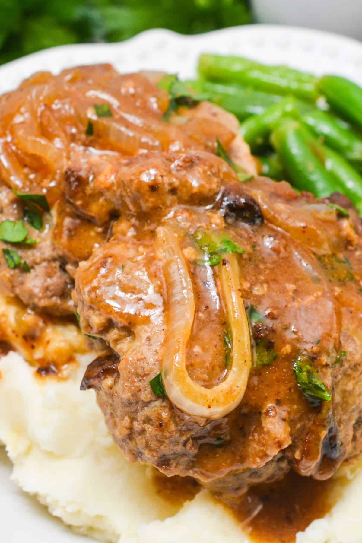 Salisbury steak with onion gravy, also known as Hamburger Steak with Onion Gravy, served over mashed potatoes and accompanied by green beans on a white plate.