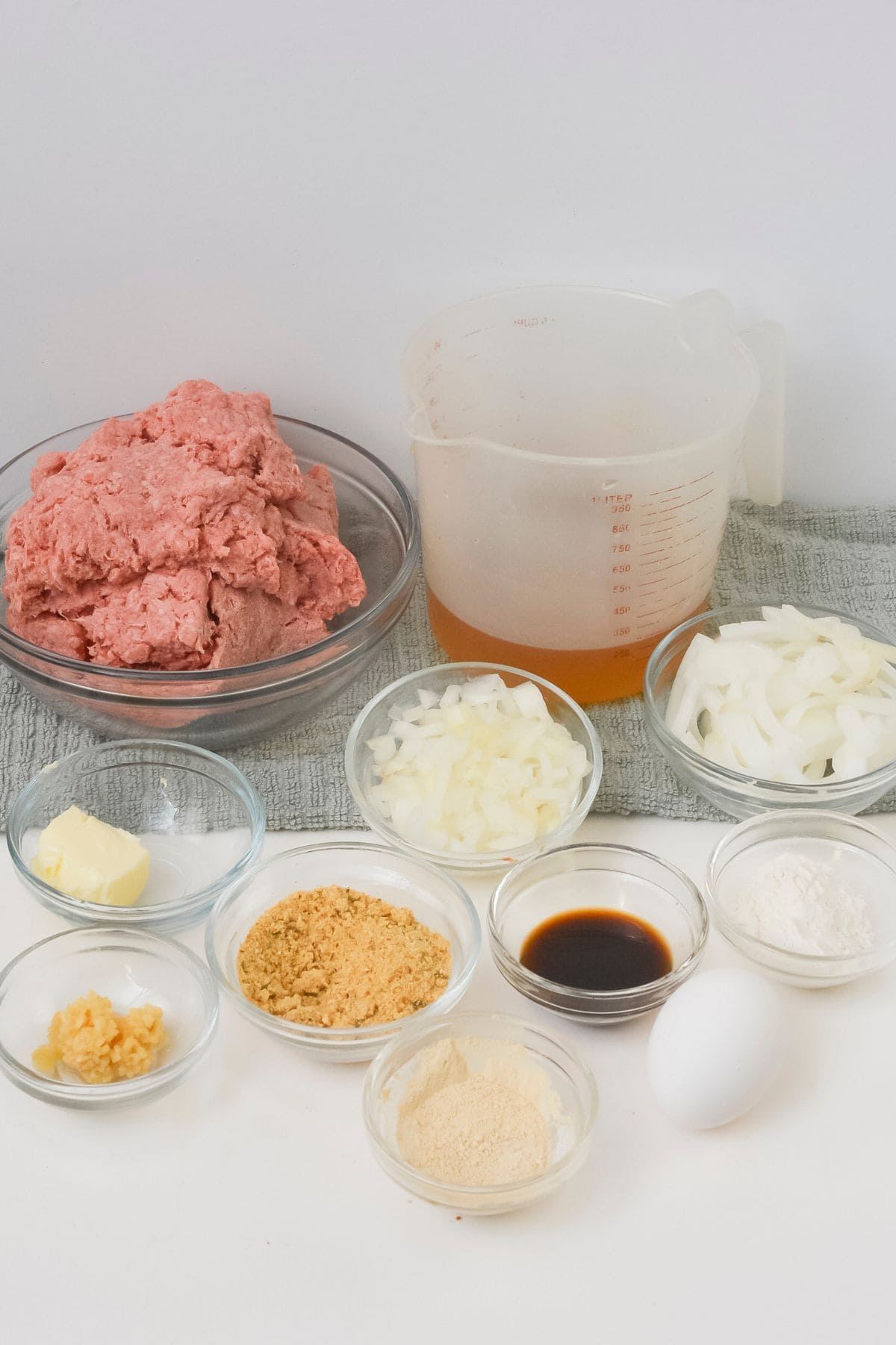 A variety of ingredients in glass bowls  including ground meat, chopped onions, butter, egg, seasonings, breadcrumbs, garlic, flour, Worcestershire sauce, and a measuring cup with broth.
