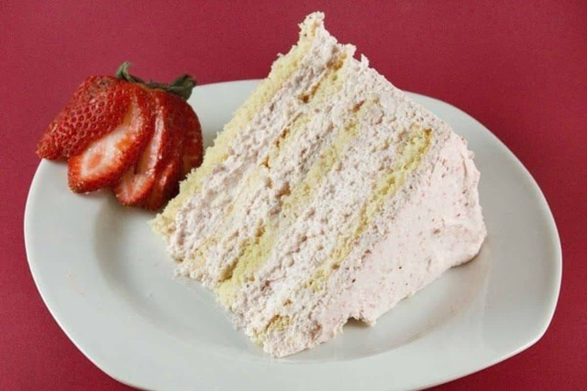 A slice of layered strawberry cake with pink frosting is served on a white plate next to three sliced strawberries, set against a red background—one of the prettiest desserts perfect for spring recipes.
