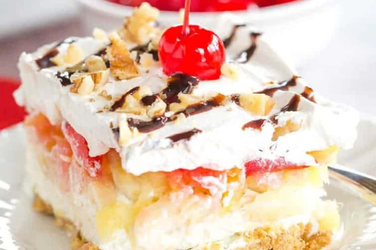 A slice of layered dessert with whipped cream, pineapple, sliced bananas, chopped nuts, chocolate drizzle, and a maraschino cherry on top—one of the prettiest desserts and perfect for your spring recipes. Served on a white plate.