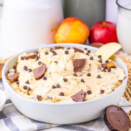 A bowl of creamy Peanut Butter Dip topped with chocolate chips and peanut butter cups, surrounded by apples and a glass of milk on a striped cloth.
