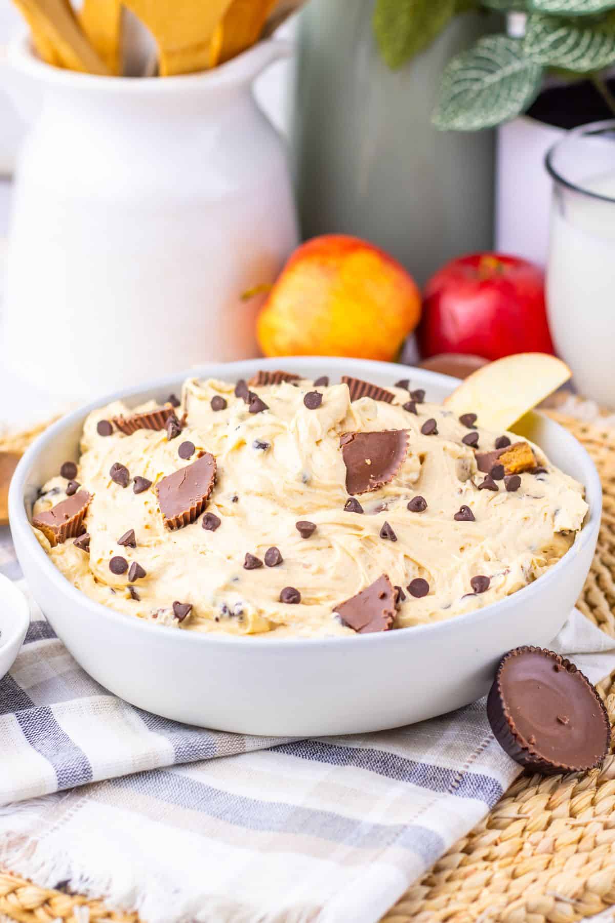 A bowl of creamy Peanut Butter Dip topped with chocolate chips and peanut butter cups, placed on a cloth with apples and a glass of milk in the background.