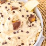 A bowl of creamy Peanut Butter Dip topped with chocolate chips and peanut butter cup pieces, with an apple slice dipped in, placed on a woven mat.