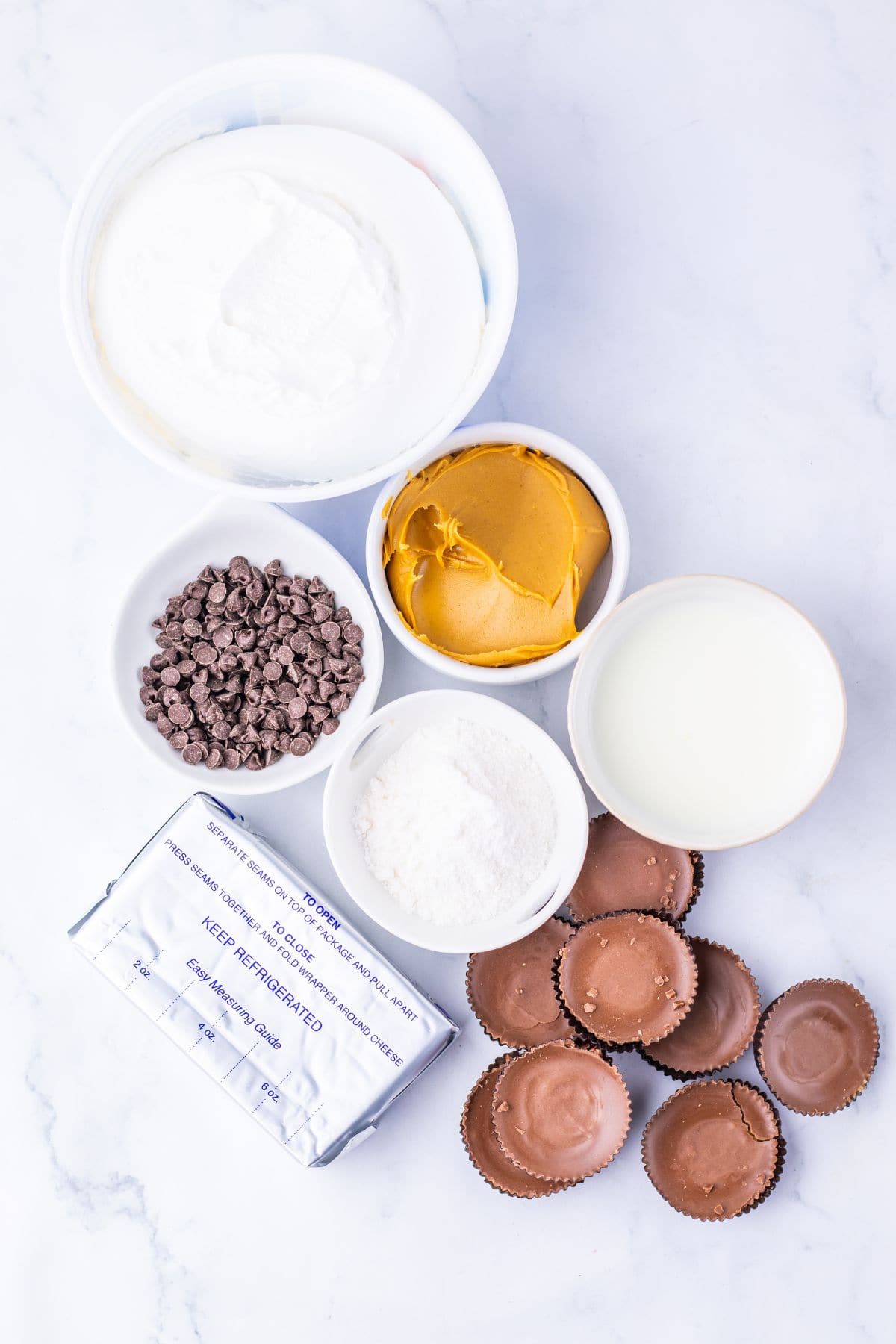 Assorted ingredients for Peanut Butter Dip, including cream cheese, chocolate chips, peanut butter, milk, powdered sugar, whipped topping, and peanut butter cups on a white surface.
