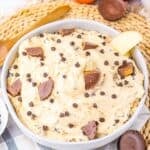 A bowl of creamy Peanut Butter Dip topped with chocolate chips and peanut butter cup pieces, surrounded by apples and snacks on a woven mat.