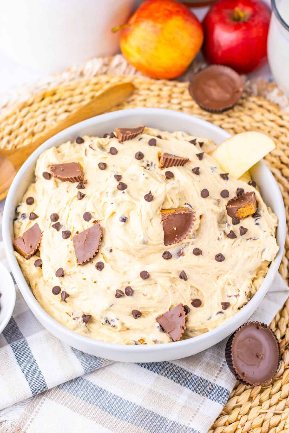 A bowl of creamy Peanut Butter Dip topped with chocolate chips and peanut butter cup pieces, surrounded by apples and snacks on a woven mat.