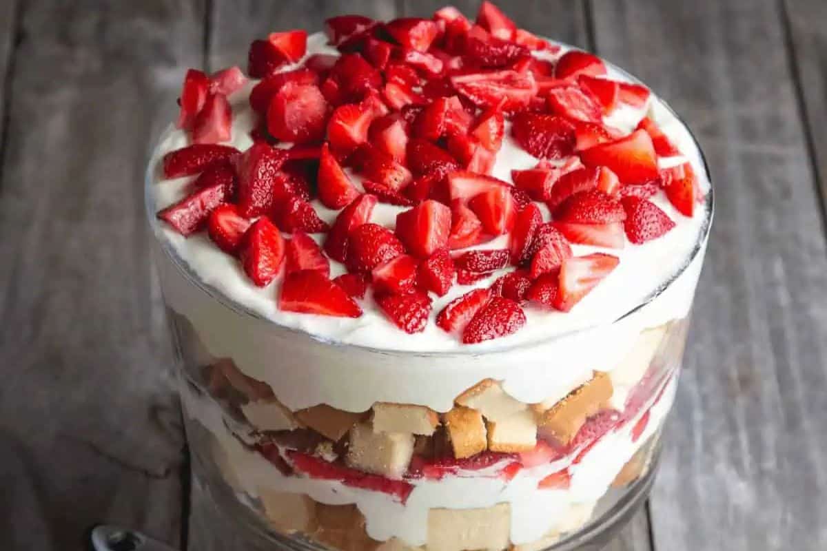 A glass trifle dish filled with layers of cake cubes, whipped cream, and sliced strawberries on a wooden surface—one of the prettiest desserts and a must-try for delightful spring recipes.