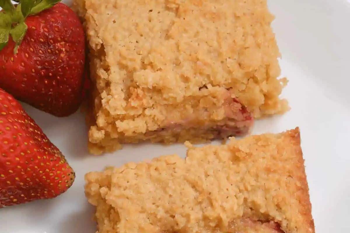 Two strawberry oat bars on a white plate next to two fresh strawberries—one of the prettiest desserts and perfect for spring recipes.