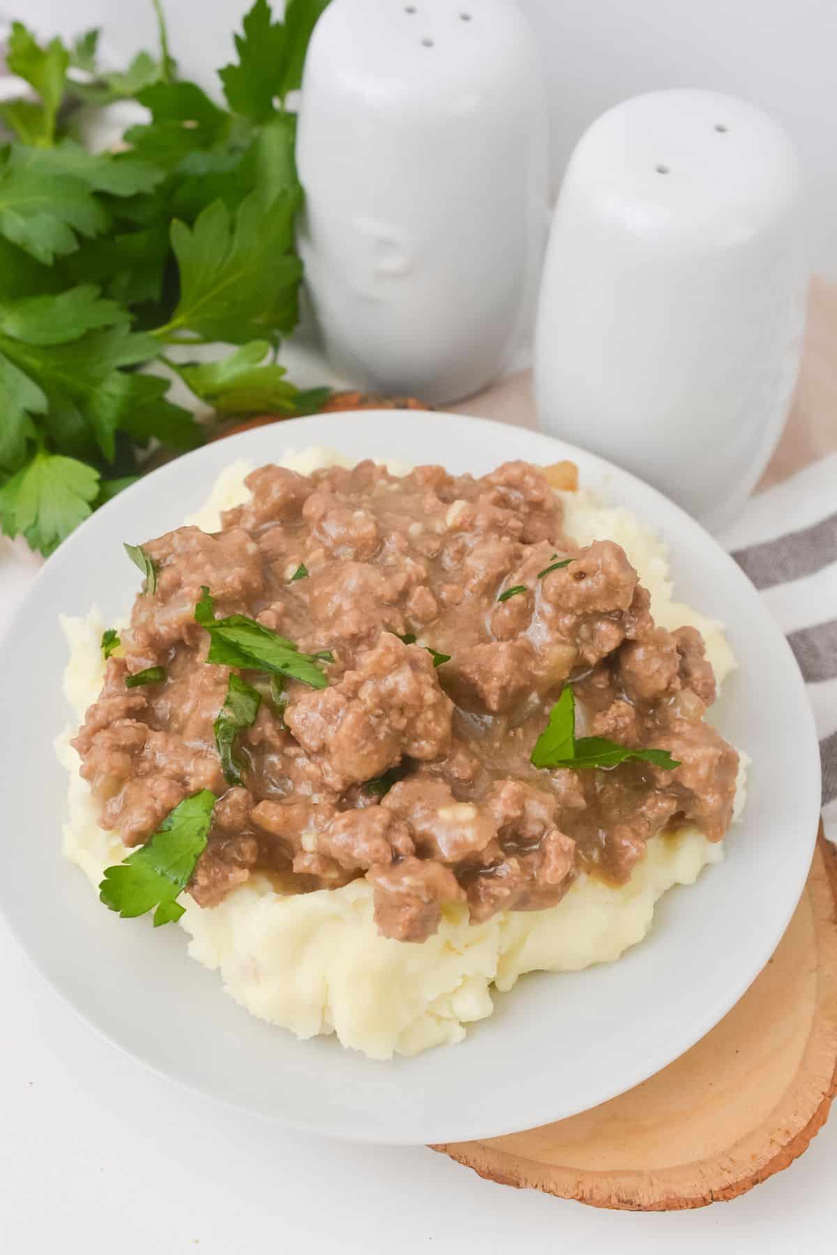 A plate of mashed potatoes topped with garnished with parsley, sits next to salt and pepper shakers and fresh parsley.
