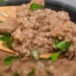 A close-up of a wooden spoon holding refried beans garnished with chopped parsley, with more beans and a hint of savory hamburger gravy visible in the background.