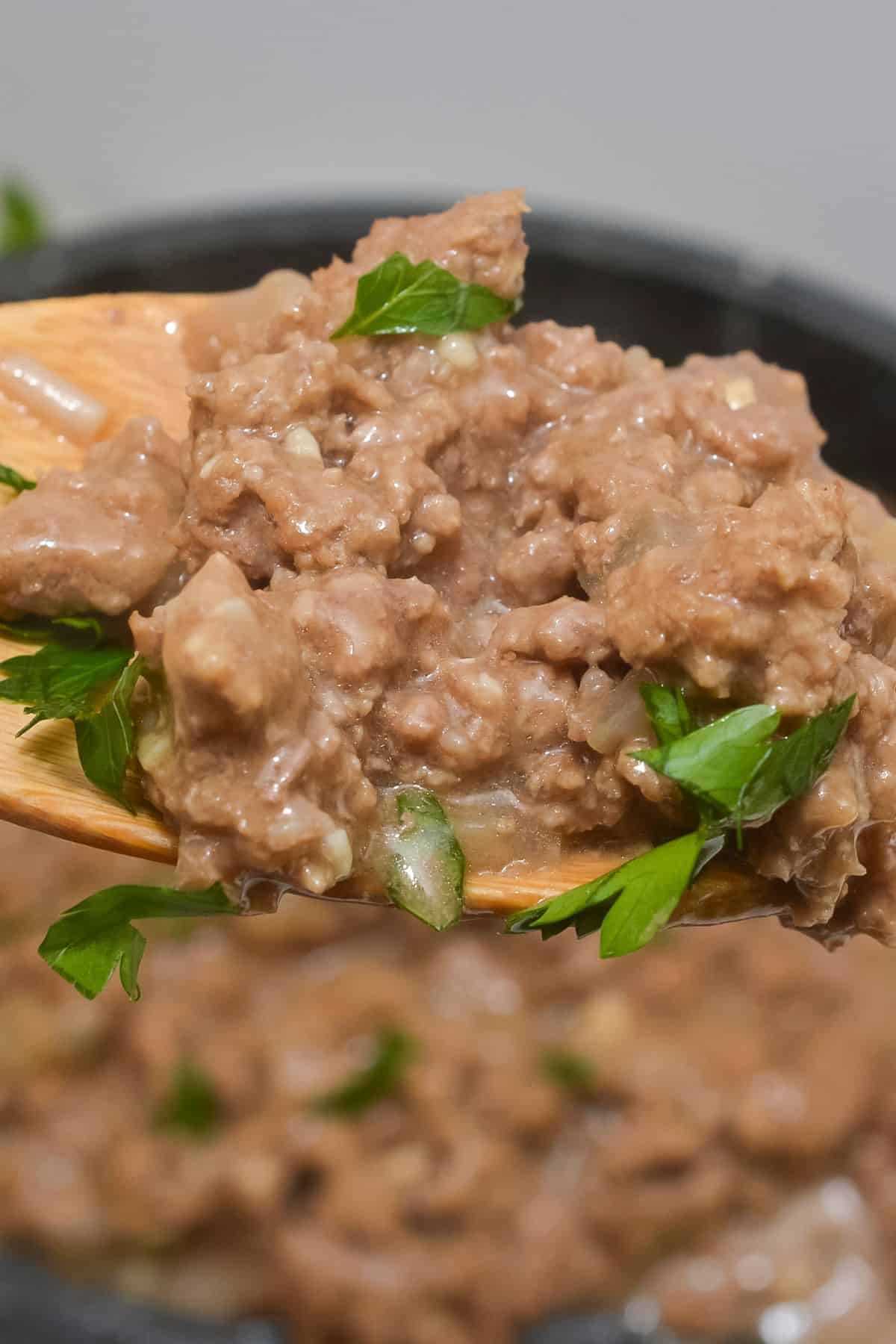 A close-up of a wooden spoon holding refried beans garnished with chopped parsley, with more beans and a hint of savory hamburger gravy visible in the background.