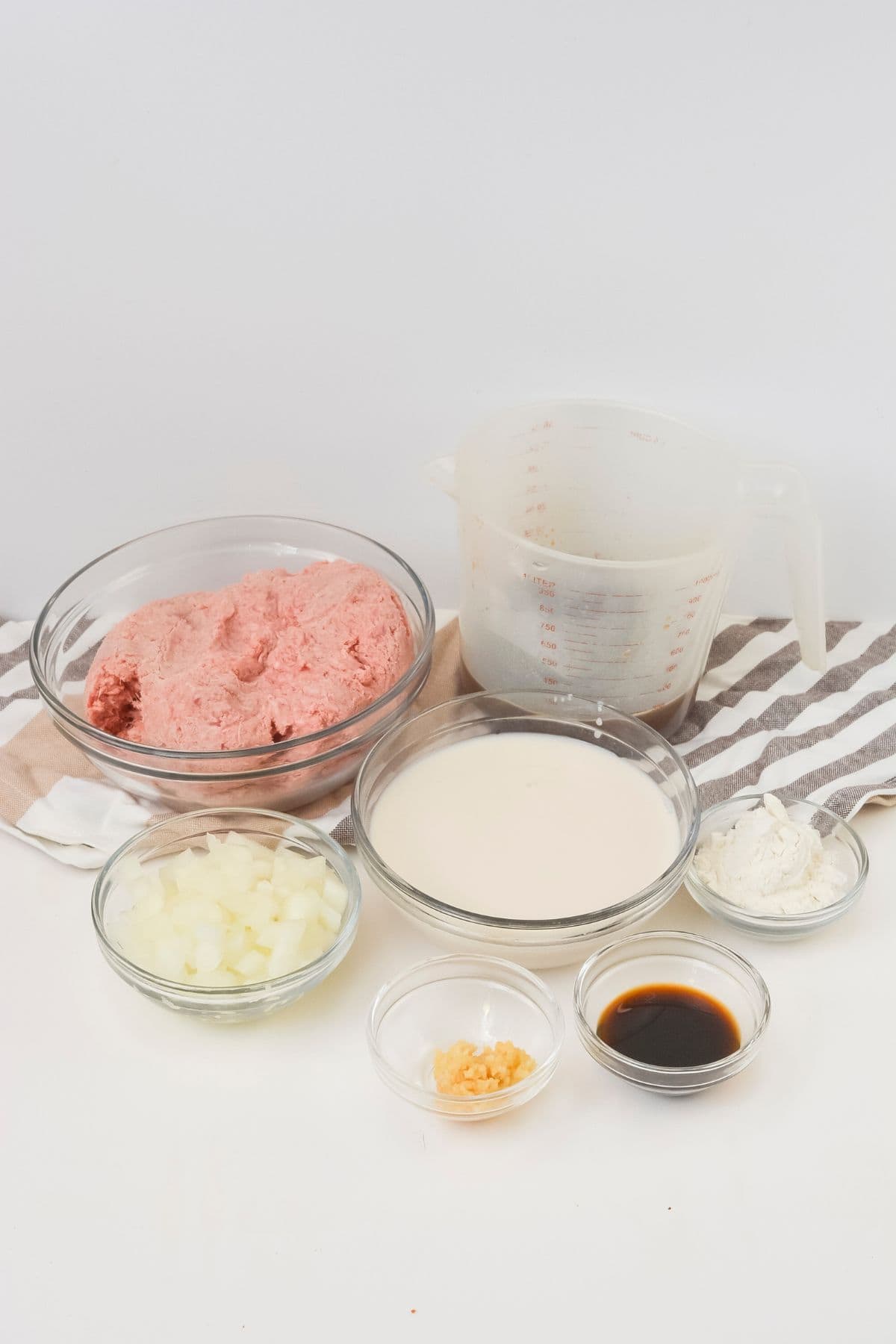 Several clear bowls containing ingredients for hamburger gravy—ground meat, chopped onions, garlic, milk, flour, soy sauce, and a measuring jug with liquid—are arranged on a striped cloth against a white background.