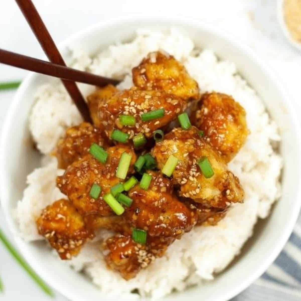 A bowl of white rice topped with saucy chicken pieces, garnished with sesame seeds and chopped green onions—this dish is truly an It's a Keeper meal, served with chopsticks resting in the bowl.