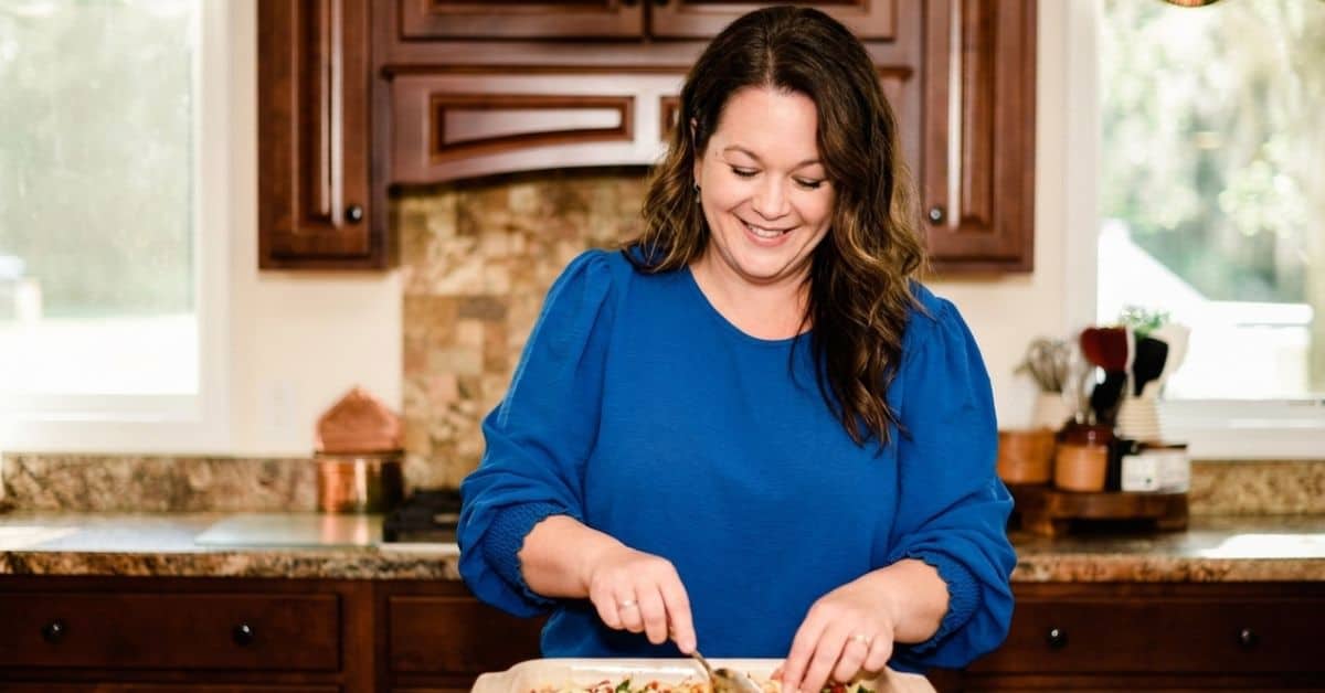 Christina, in a blue blouse, stands in the kitchen, smiling warmly as she prepares food on a baking tray.