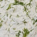 A bowl of creamy Dill Dip topped with chopped fresh dill sits on a striped cloth, surrounded by potato chips and sprigs of dill.